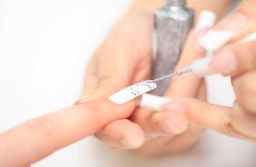 Person applying silver nail polish on fingernail with white base coat.