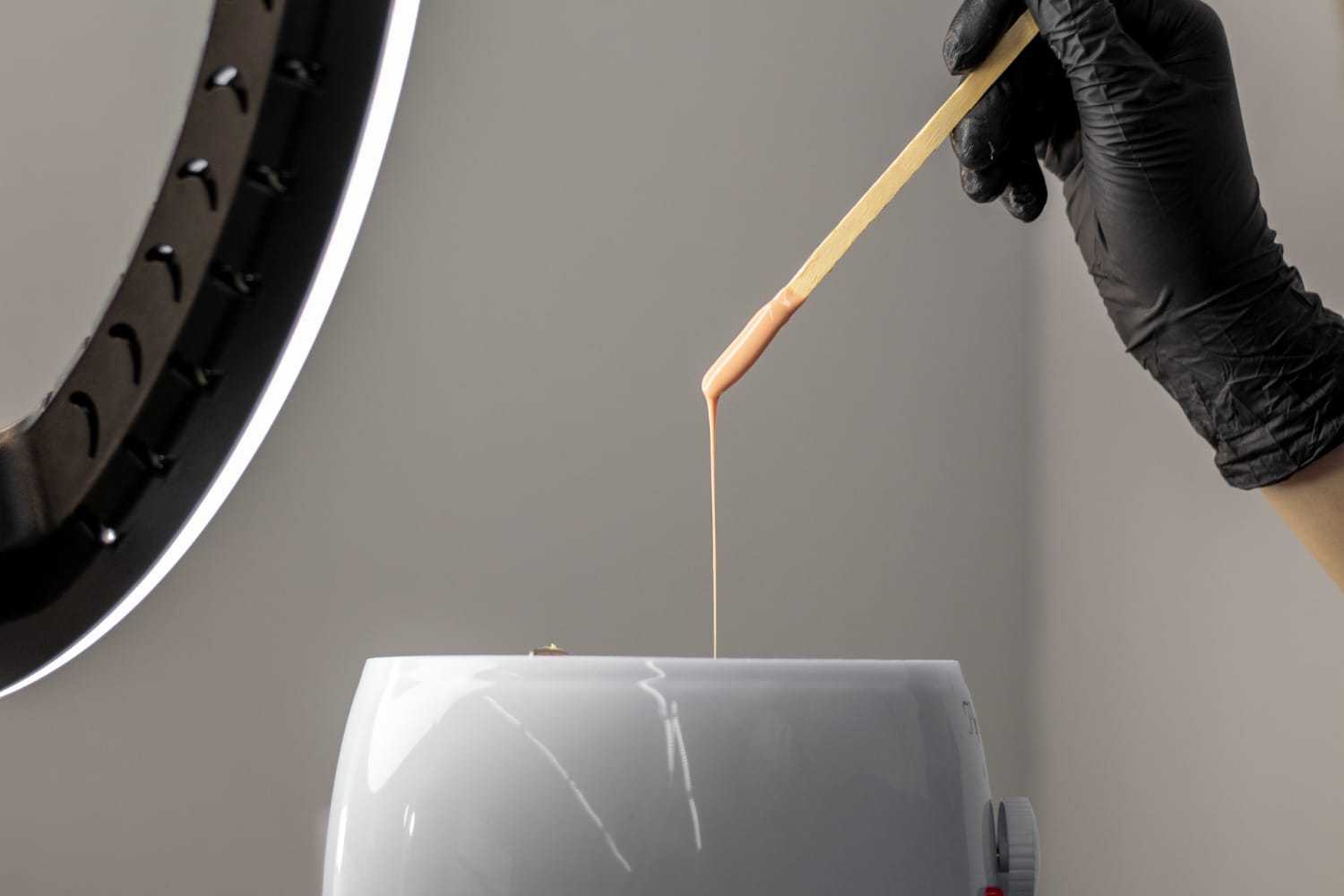 Gloved hand holding a spatula with warming wax over a wax heater.