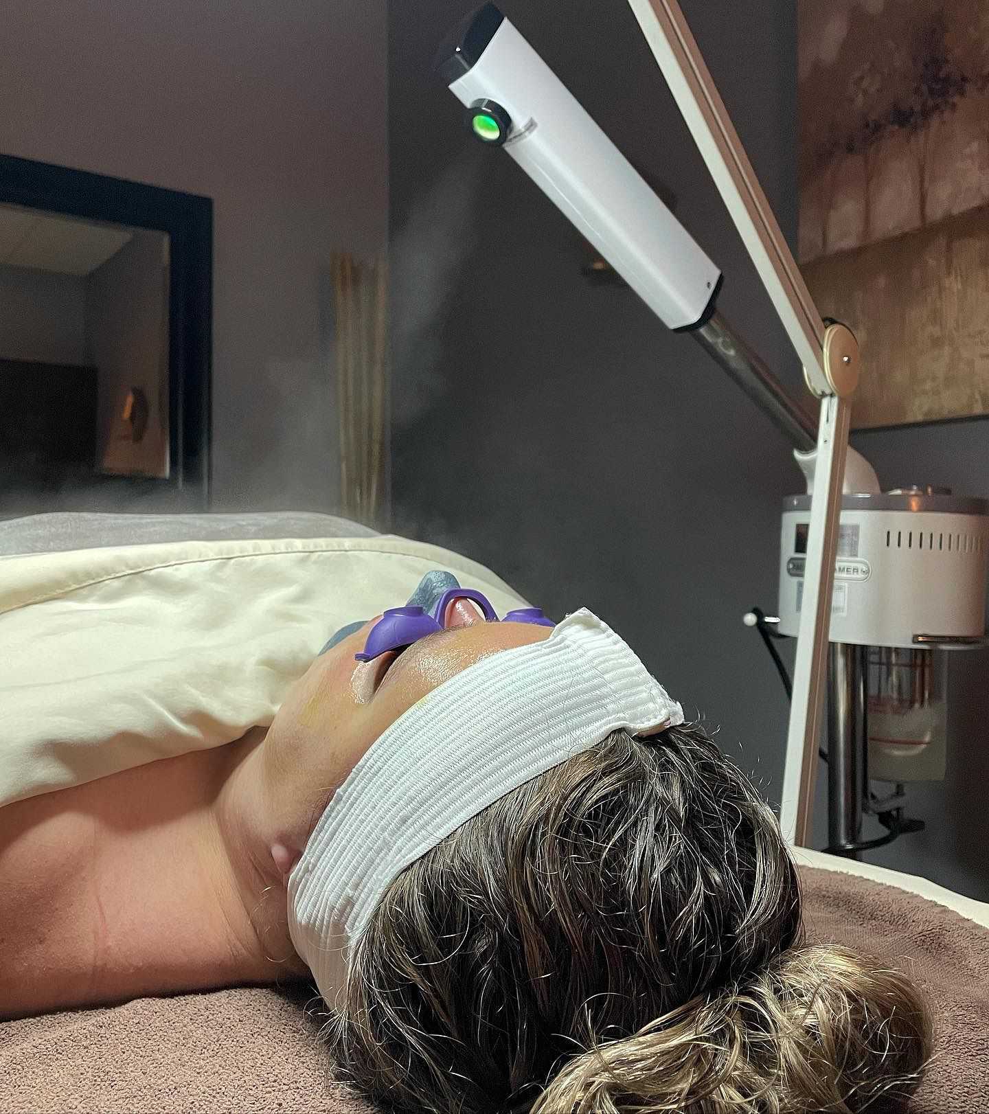 Person receiving a facial steam treatment at a spa, lying on a table with a headband and towel.