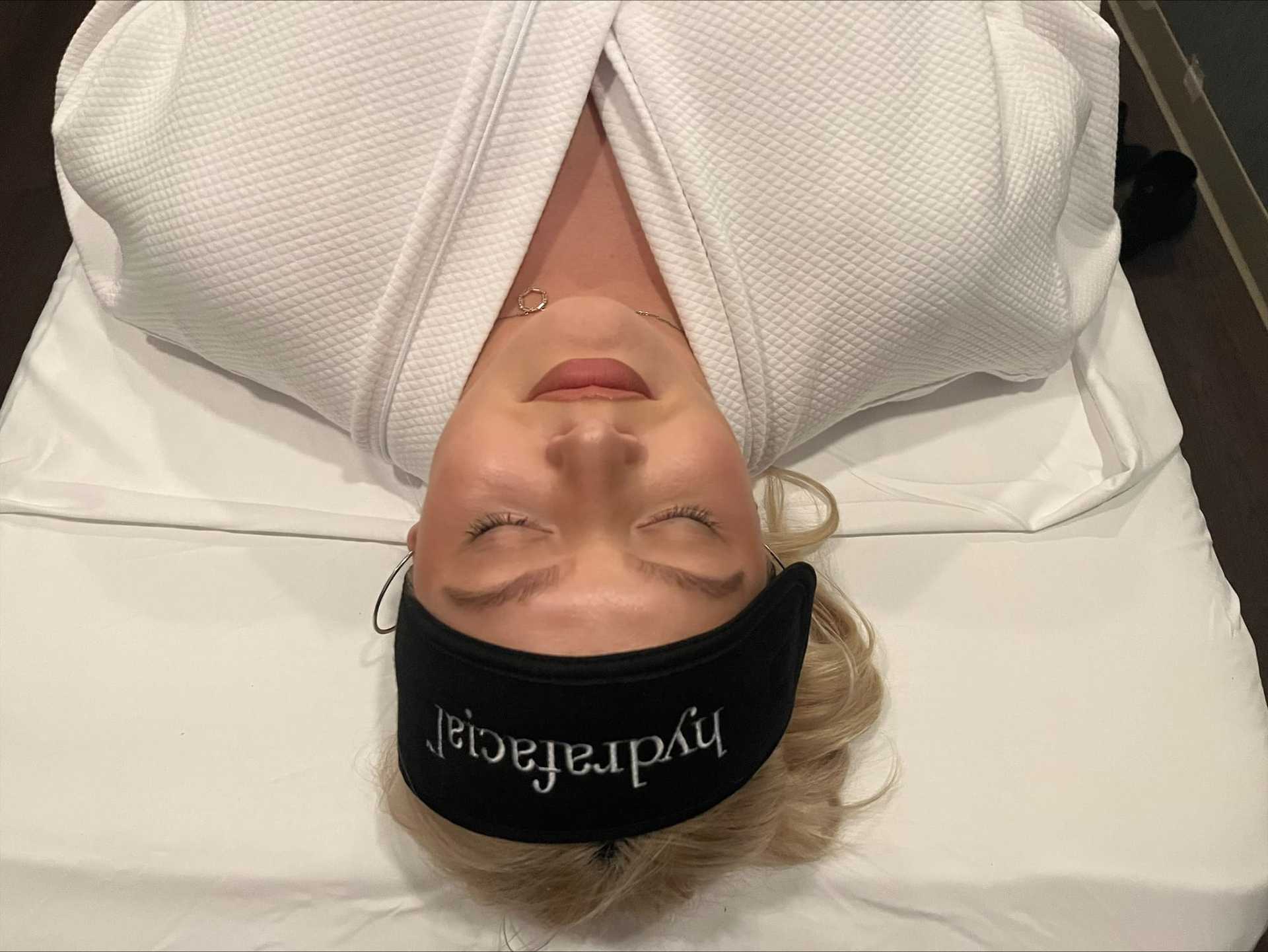 Person with a black headband and white robe relaxing on a spa bed with eyes closed.