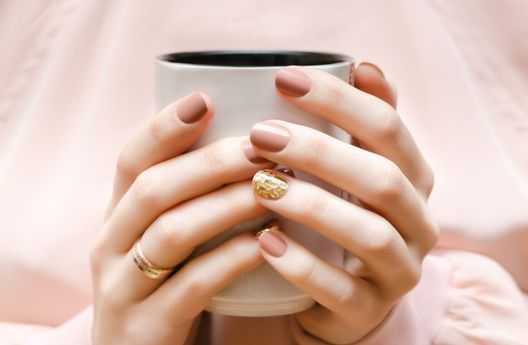 Hands with mauve nail polish holding a white mug, showcasing an intricate gold design on one nail.