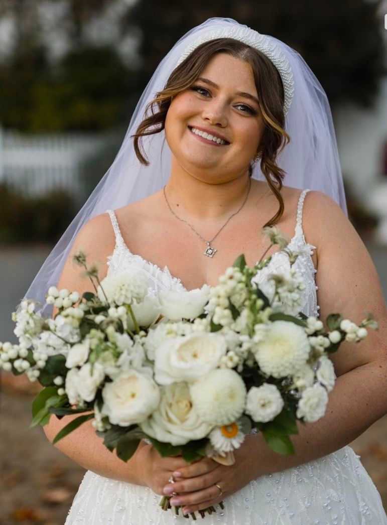 Bride with bouquet of white flowers smiling outside in a veil and white wedding dress.
