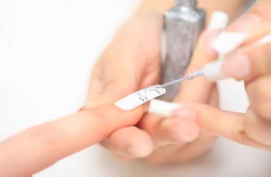 Person applying silver nail polish on fingernail with white base coat.