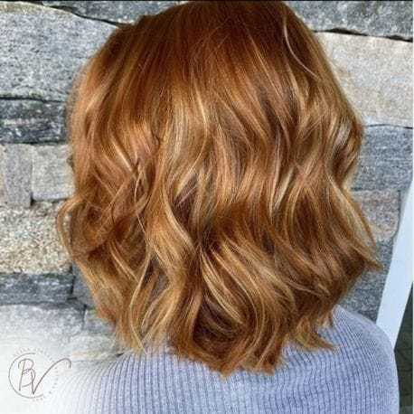 Woman with shoulder-length wavy ginger hair against a stone wall background.