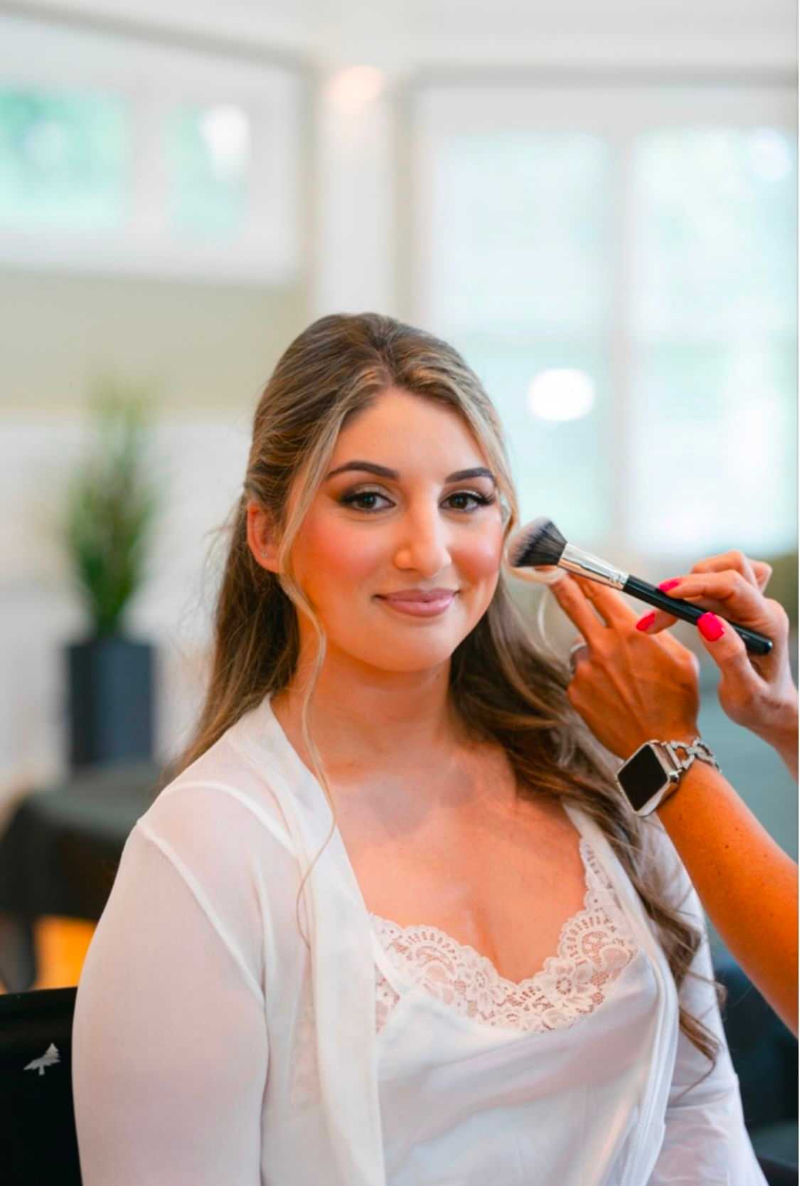 Woman getting makeup applied with a brush, wearing a white lace top, and smiling softly.