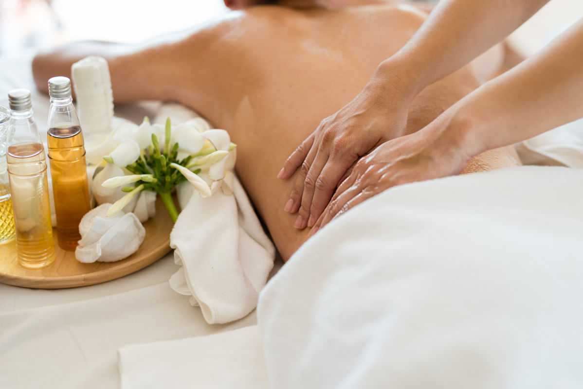 Person receiving a back massage with aromatic oils and fresh flowers on a white bed.